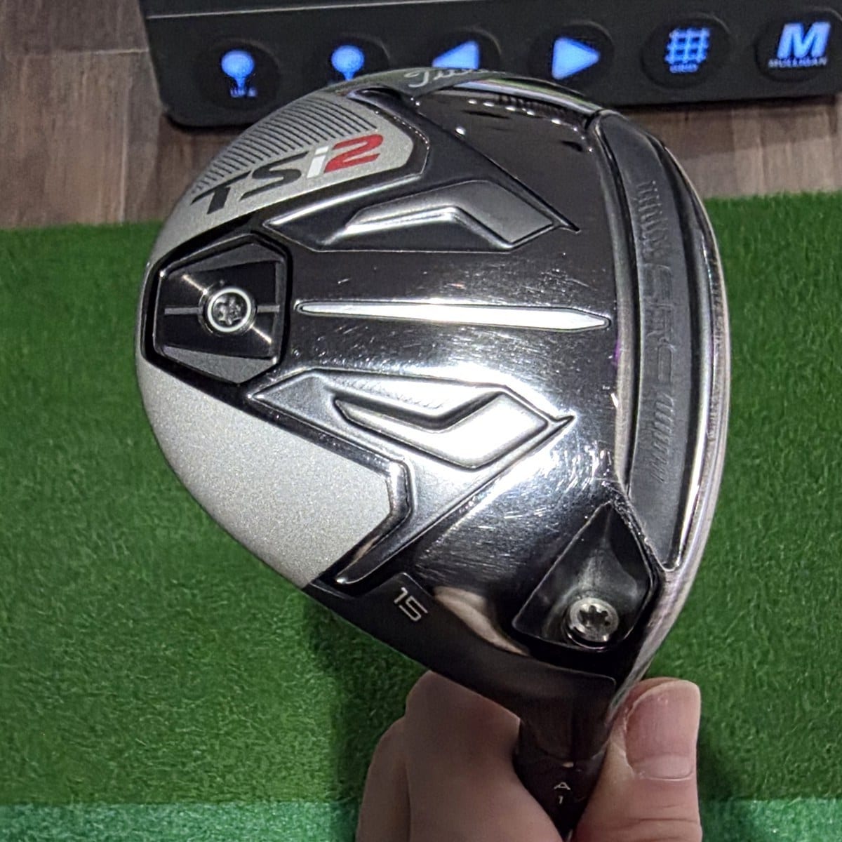 The sole of a Titleist TSi2 3-wood held up by the hosel in the author's hand, with the TSi2 badge at the top left and the loft number 15 engraved near the hosel, in front of a simulator mat and control box.