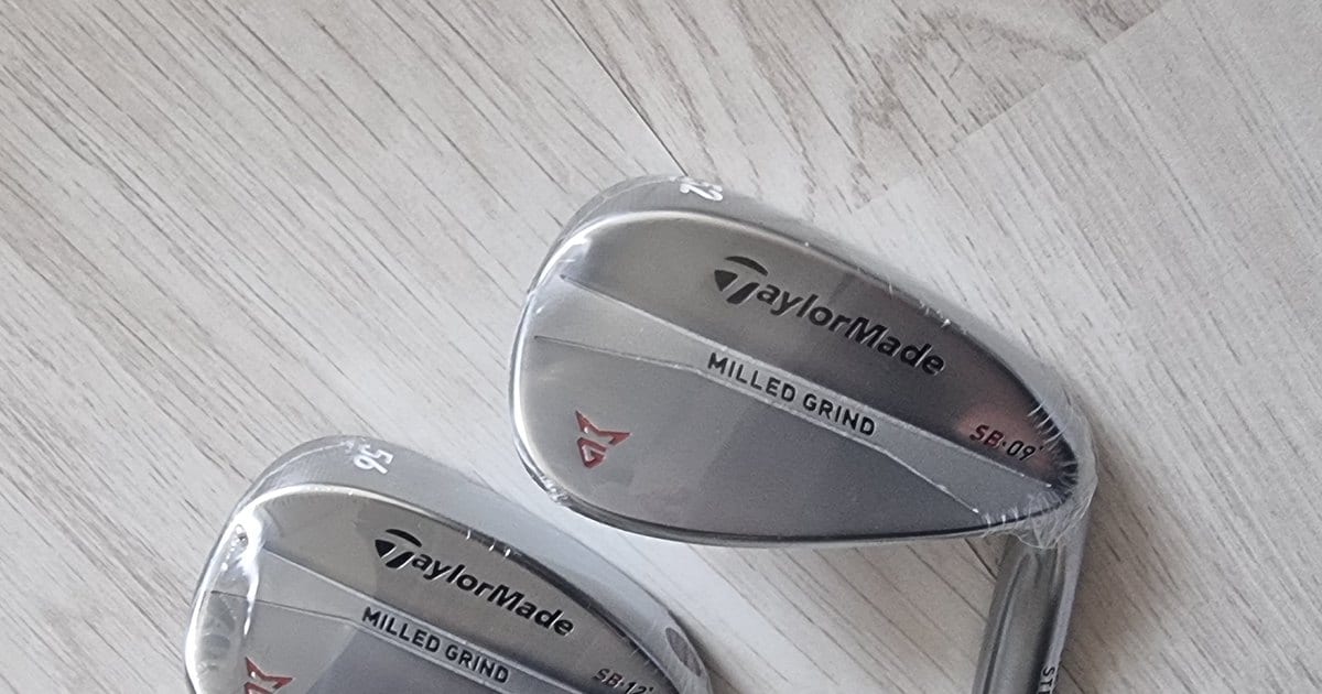 Two TaylorMade Milled Grind wedges still in their wrapping, the pair as it's usually sold, the loft stamps reading 52 and 56 on the top edge of each head.