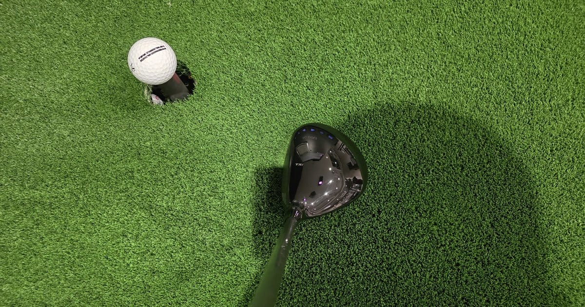 A teed golf ball and a Titleist driver head resting on the green mat of an indoor screen-golf bay.