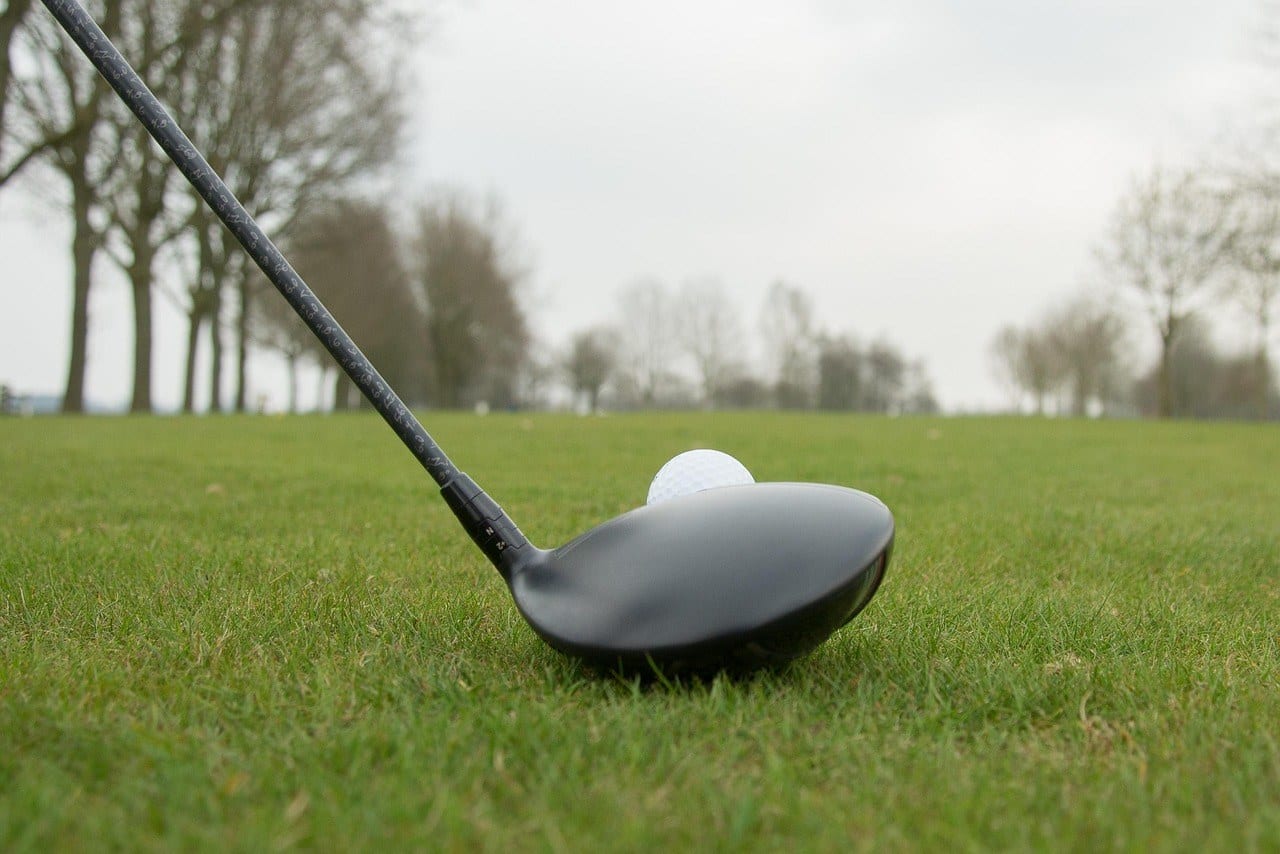 A red golf ball on a tee directly in front of a driver's face, the club blurred behind it against a dark background.