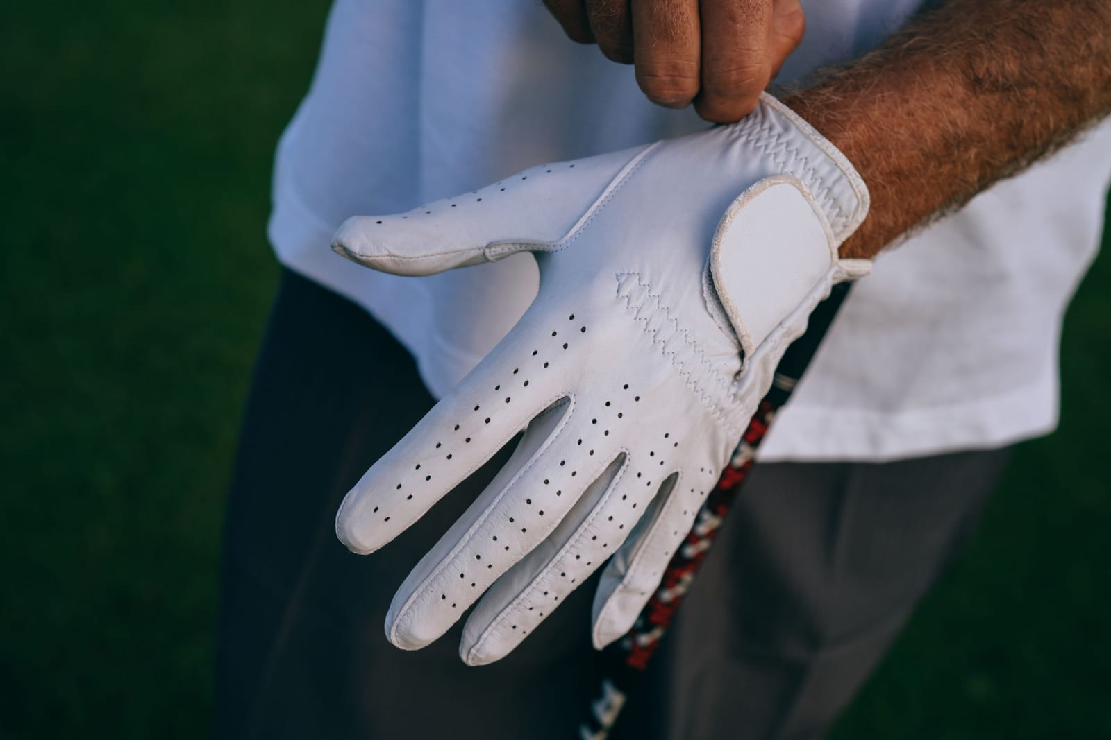 A golfer pulling a smooth white golf glove onto the left hand by the wrist strap, with neat rows of small holes across the fingers and a club grip resting against the leg.