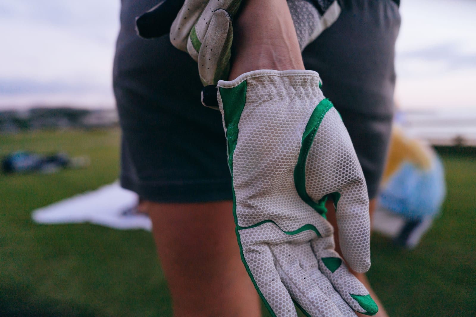 A golfer at a driving range pulling on a white golf glove with green trim, its palm and fingers covered in a raised honeycomb texture, with a second glove held in the other hand and green grass behind.