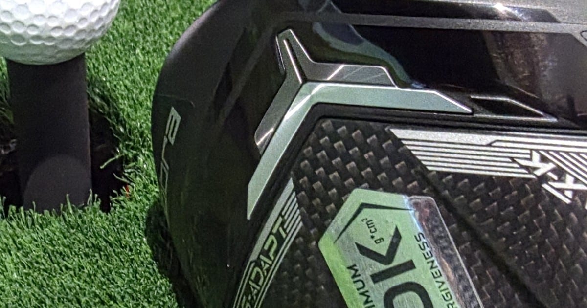 A close-up of the author's Cobra DS-ADAPT driver crown resting on artificial turf, with a teed ball behind it and a 10K MAXIMUM FORGIVENESS badge on the carbon weave.