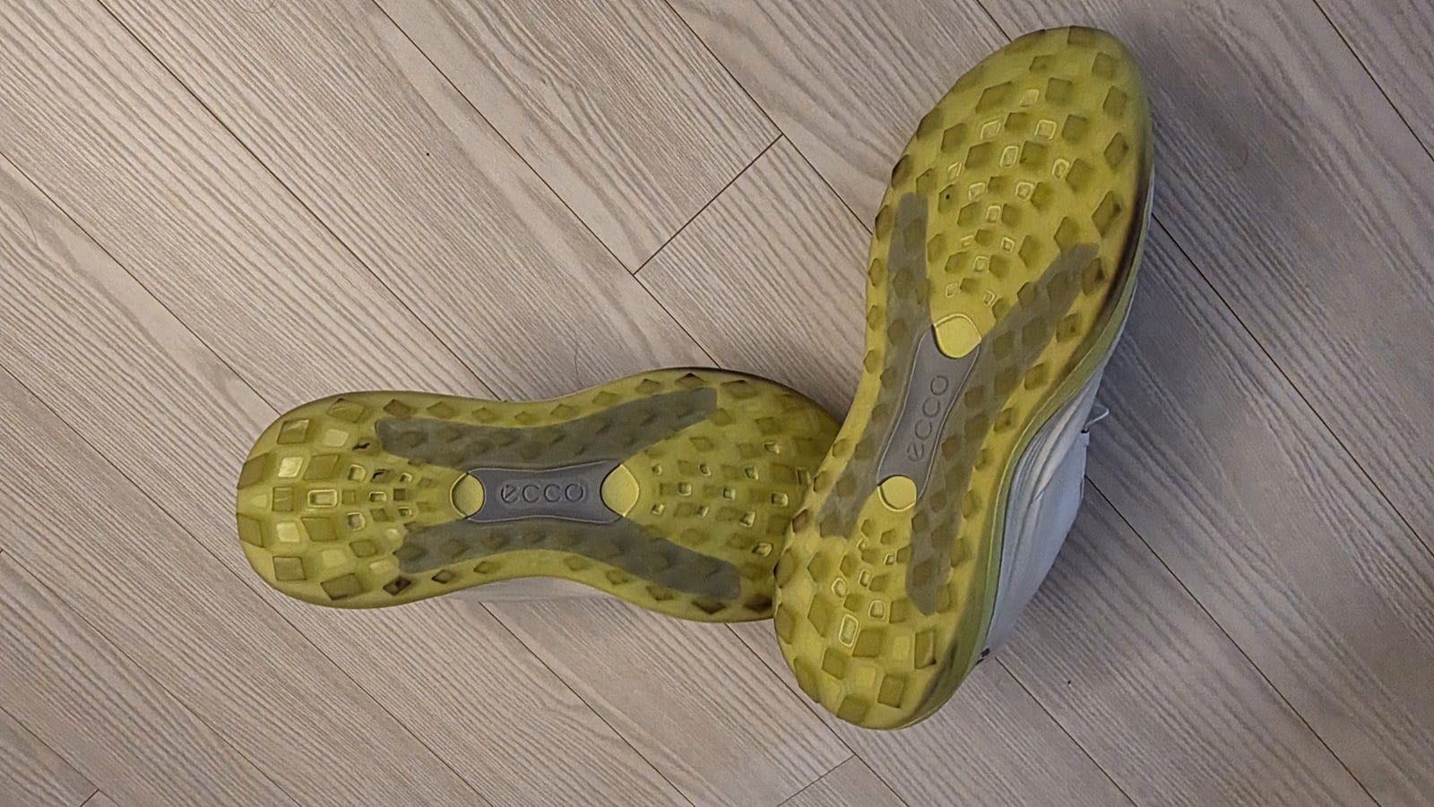 The soles of two white ECCO LT1 golf shoes on a light wood floor, showing lime-green rubber lugs, the ECCO logo on a grey panel under the arch, and darker scuffed patches toward the edges.