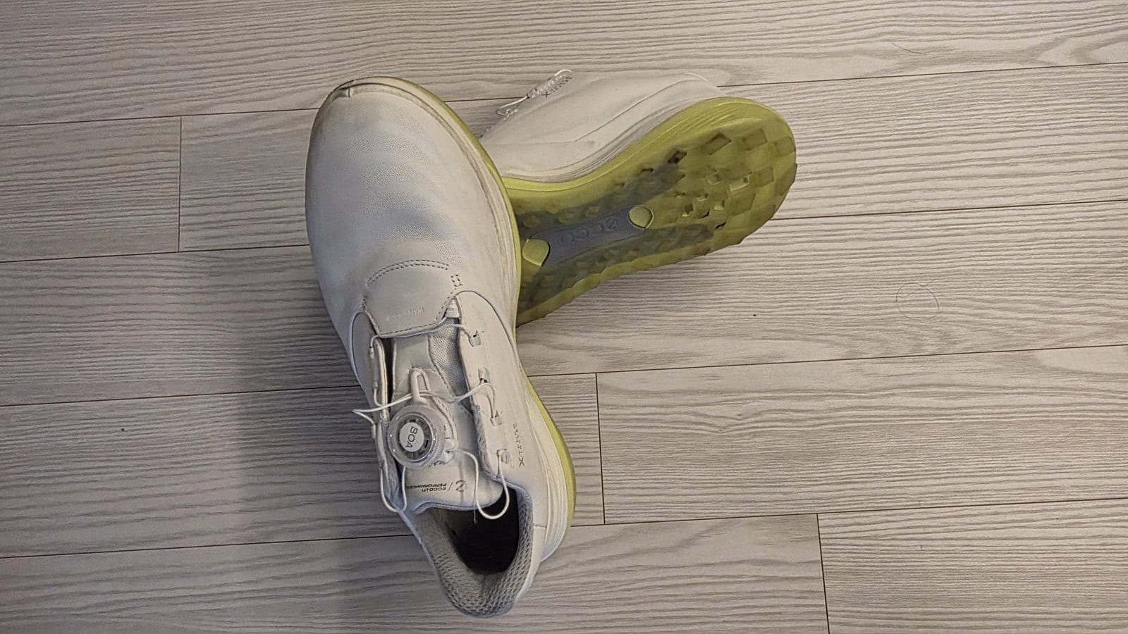 A white ECCO LT1 BOA golf shoe seen from above with its round BOA dial on the tongue, next to a second shoe tipped on its side to show the lime-green outsole, on a light wood floor.