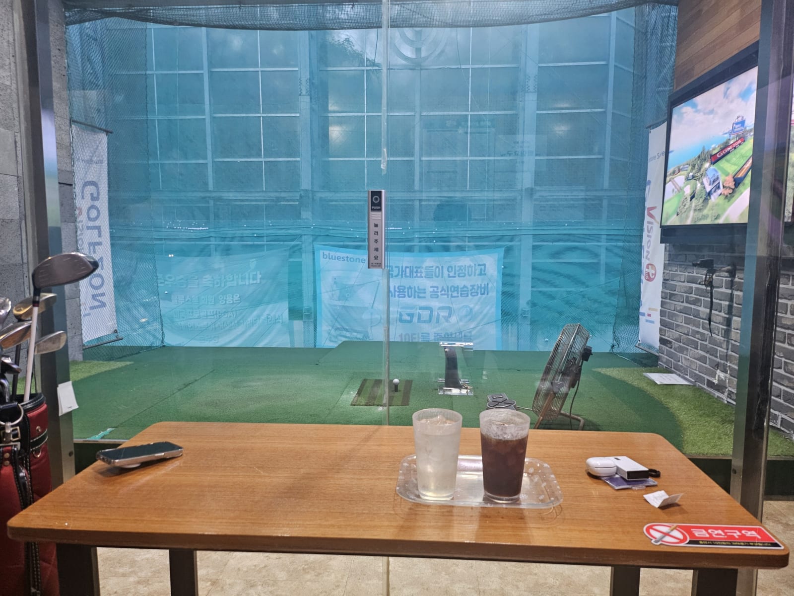 An empty indoor practice bay photographed from the drinks table behind it: a ball sitting on the hitting mat, a net across the far wall carrying a Golfzon GDR banner, and a wall screen showing a course view.