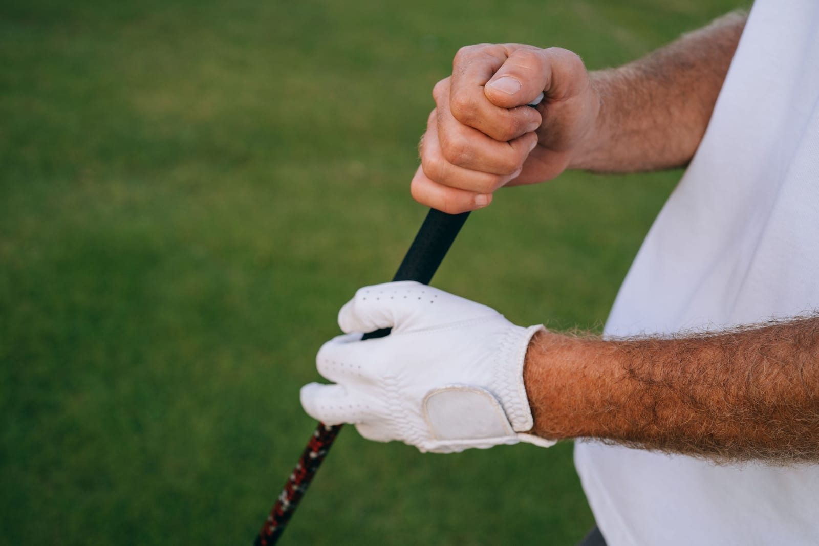 A golfer's gloved hands hold a golf club grip in a close-up on a green course, showing where the thumb and fingers press into the grip.
