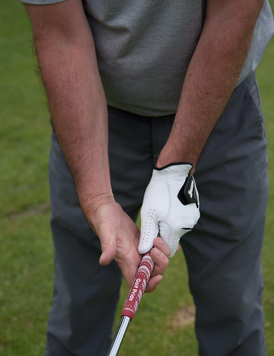 A golfer at address with both hands on a red Golf Pride grip, the white-gloved upper hand sitting above the bare lower hand, which hides the gloved fingertips from view.