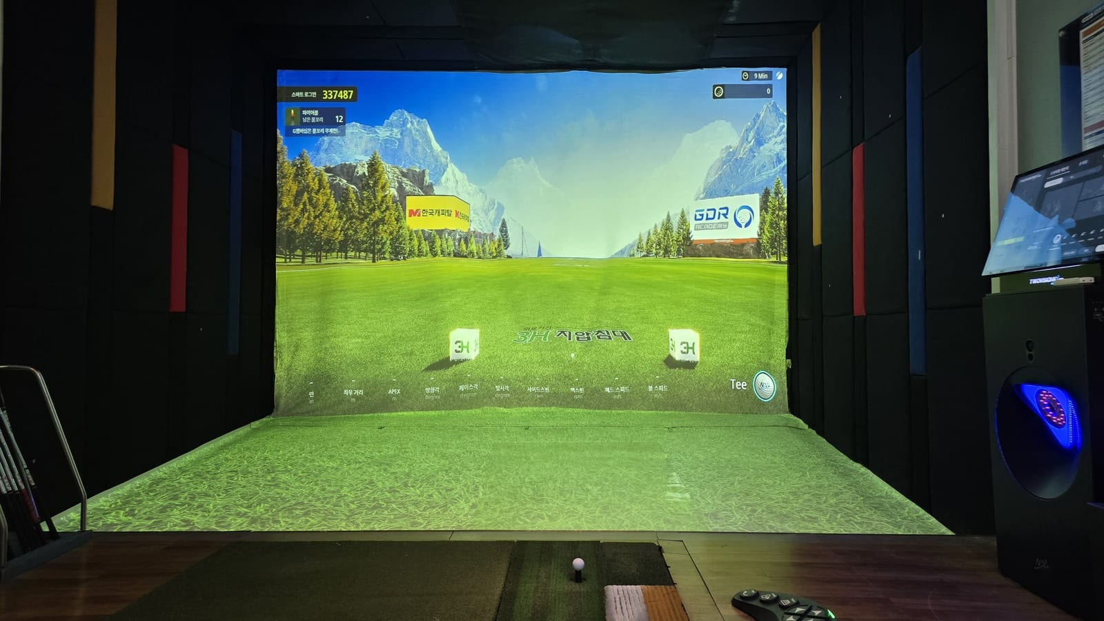A Korean screen golf bay seen from behind the tee, with a wide projection screen showing a fairway framed by mountains, a green hitting mat and a ball on a tee in the foreground, black padded walls on both sides, and a touchscreen kiosk standing to the right.