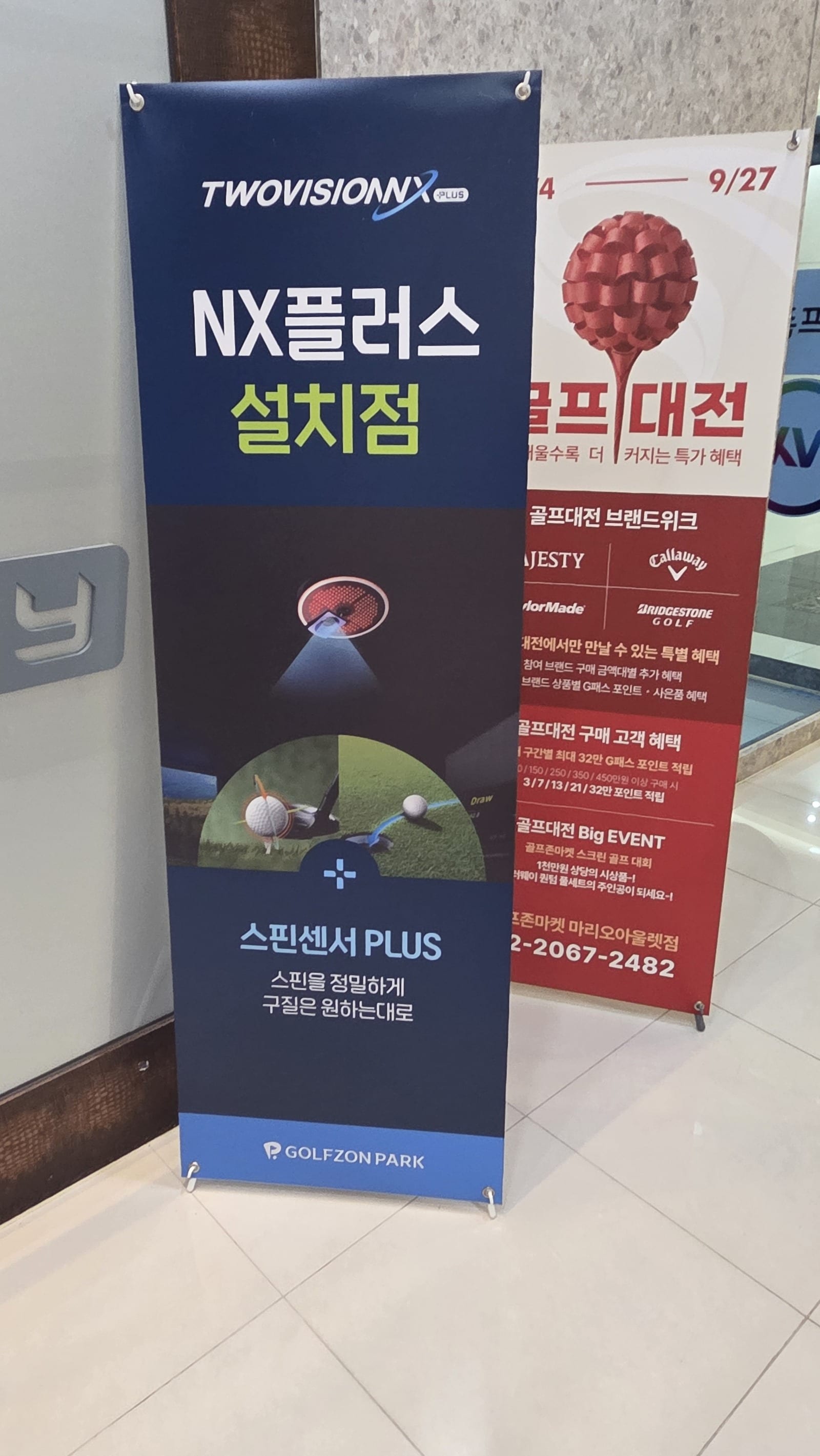 A tall navy roll-up banner standing on a tiled lobby floor. The top carries the TWOVISIONNX PLUS wordmark, below it large Korean text reading NX플러스 설치점, then an illustration of a ceiling module beaming light down onto a ball, then 스핀센서 PLUS, and the GOLFZON PARK logo on a blue strip along the bottom.