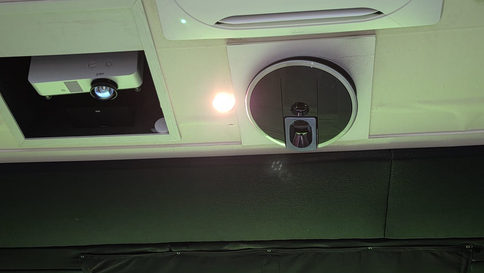 The ceiling of a screen golf booth, showing a recessed white projector on the left with its lens pointing down, and to the right a round cut-out in the ceiling panel holding a dark rectangular module with two visible lenses.