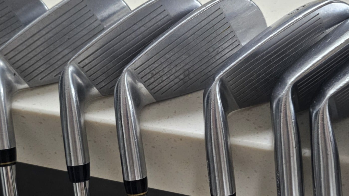 Macro photograph of a row of iron heads resting against a counter edge, with the hosels and necks in sharp focus and the grooved faces behind them.