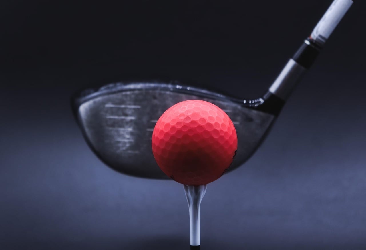 A driver head behind a red golf ball on a tee, shot against a dark background, with the lower shaft visible above the head.