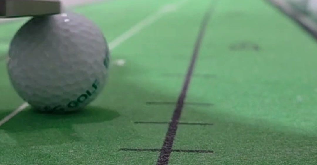 A golf ball resting beside the printed target line of an indoor putting mat, the line and its tick marks running away from the camera.