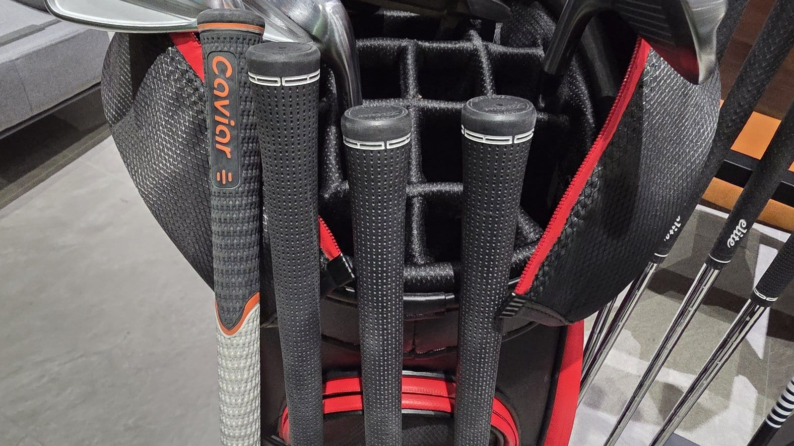 My golf bag's club well from above, showing a white and orange CAVIAR grip on the leftmost driver shaft, an Elite grip beside it, and two more Elite grips on irons behind it.