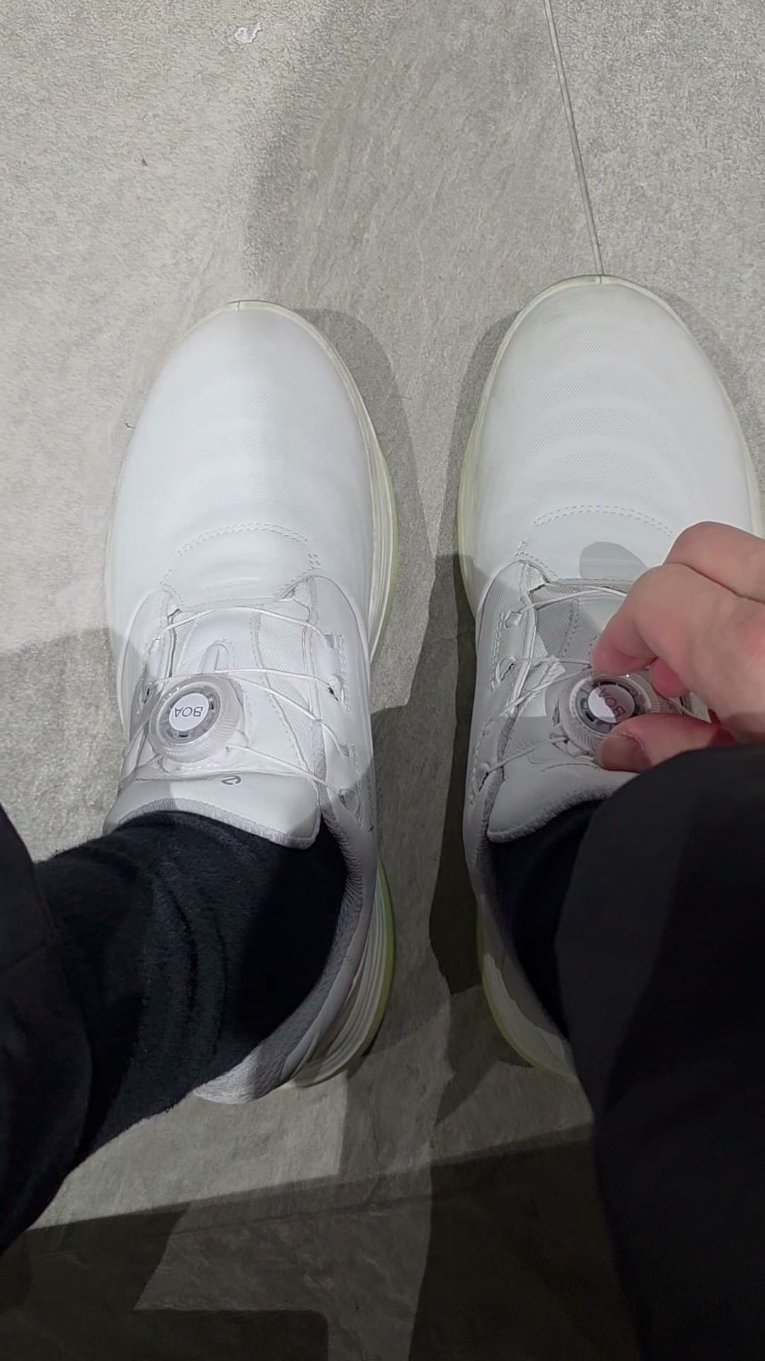 Looking down at a pair of white spikeless golf shoes with round BOA dials on the tongue, one hand turning the dial on the right shoe to tighten it, on a grey tiled floor.