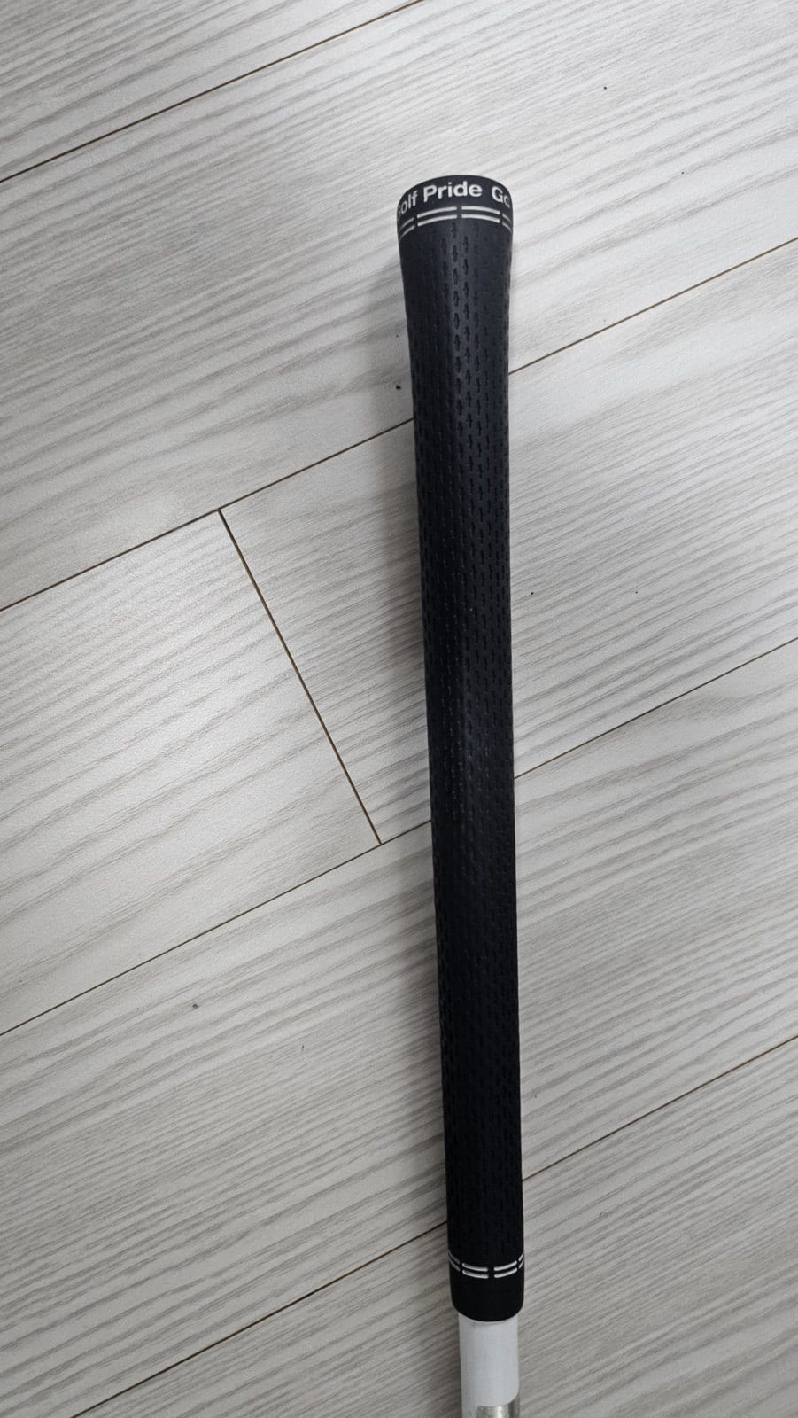 The butt end of a golf club on a pale wood floor, a black Golf Pride grip running the length of the frame with the shaft just visible below it.