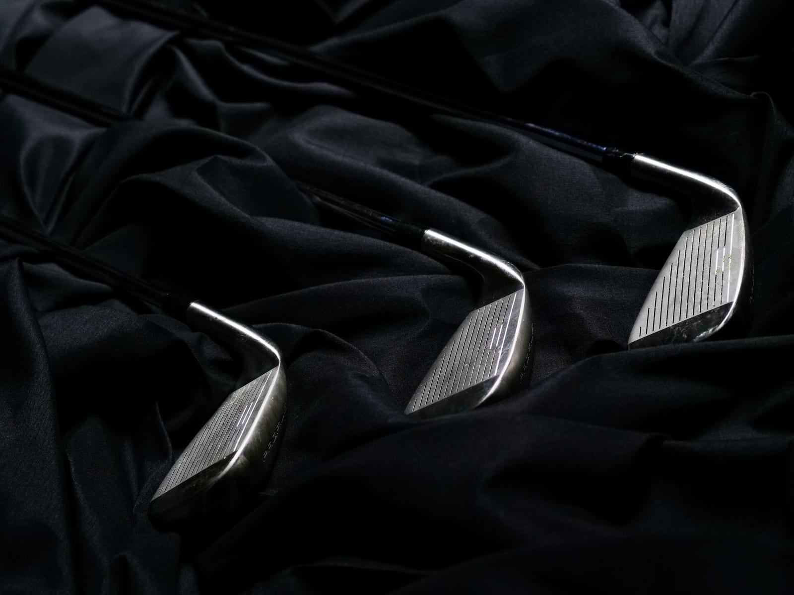 Three iron heads on dark rumpled cloth, faces toward the camera catching the only light, their shafts receding into shadow.