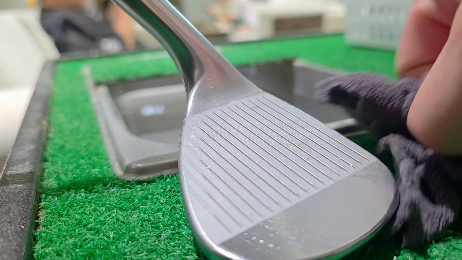A wedge face resting on a green practice mat, hosel and the lower few inches of steel shaft visible at the top, a gloved hand at the right edge of the frame.