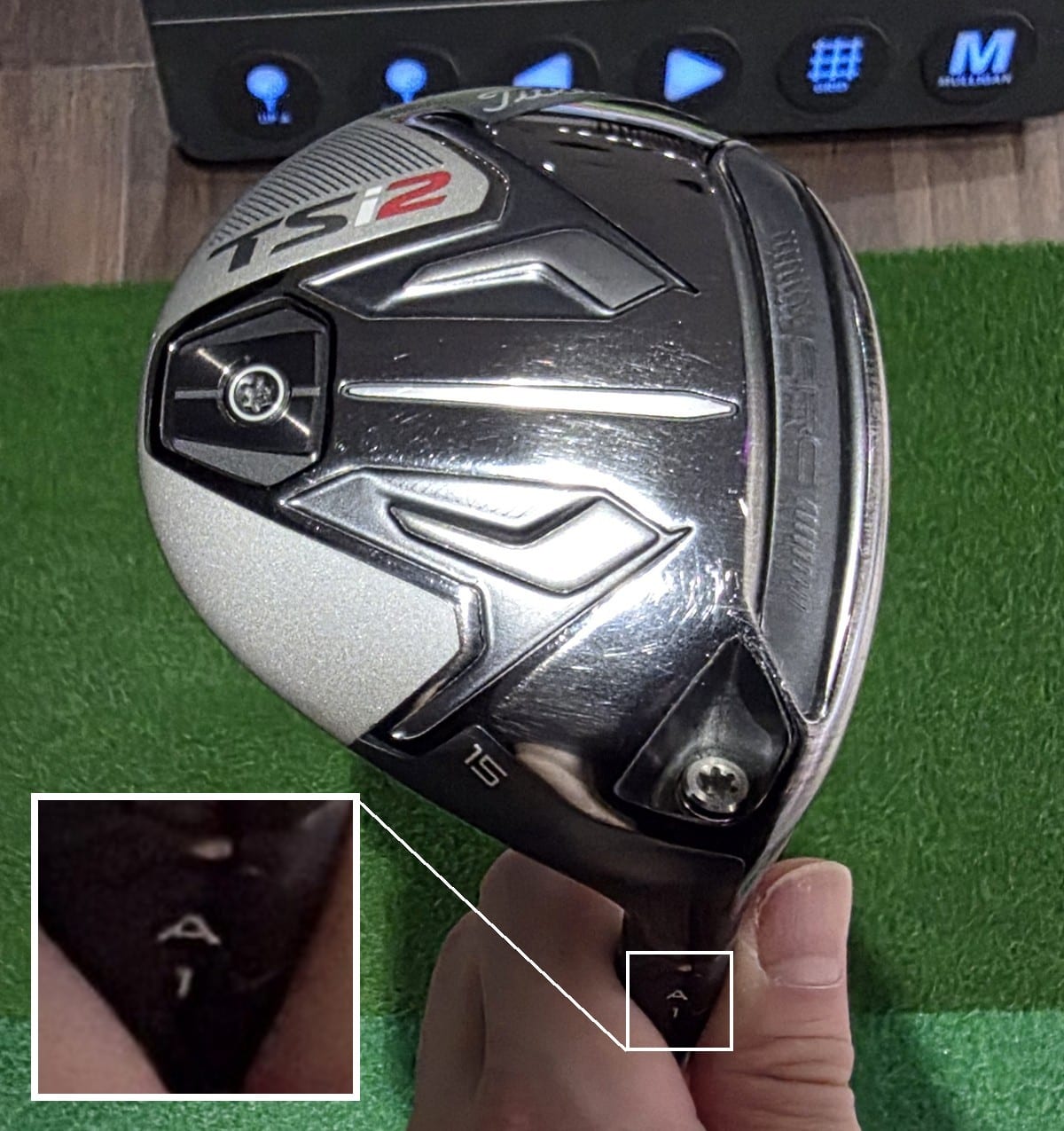 A Titleist TSi2 fairway metal held sole-up over a mat in an indoor bay. The sole carries the TSi2 badge, a 15 loft stamp and a SureFit weight screw. A boxed inset at lower left magnifies the hosel sleeve, where the letter A sits above the number 1.