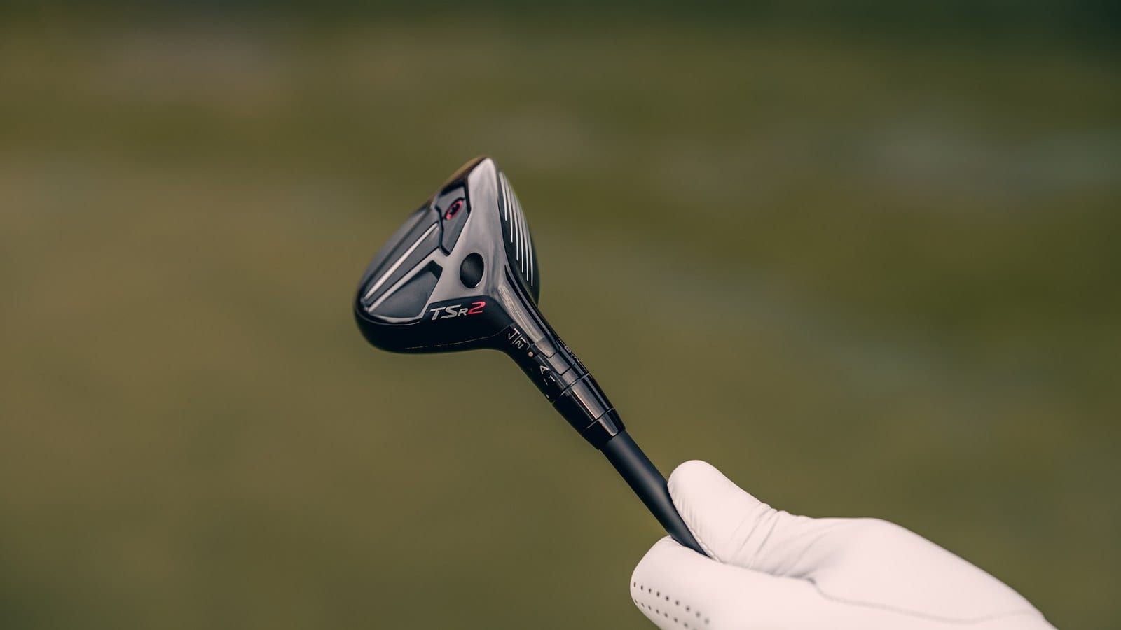 A gloved hand holds a Titleist TSR2 hybrid up against a blurred green background, with the hosel and its SureFit sleeve in view.