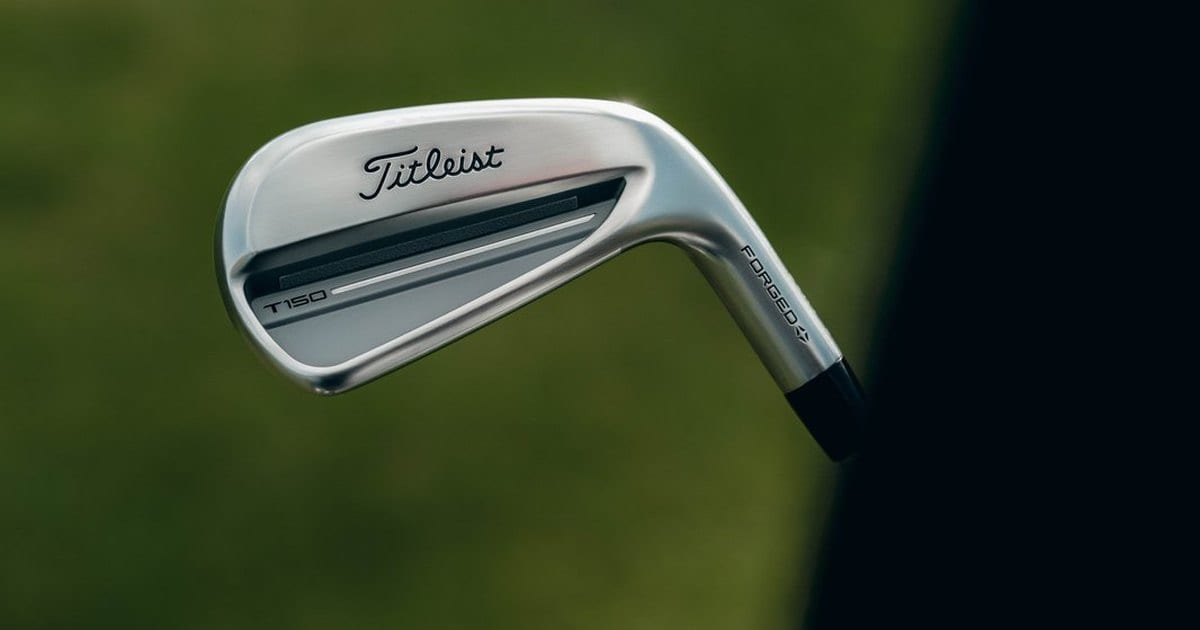 The back cavity of a 2025 Titleist T150 iron, with the T150 name stamped on the cavity bar, held against out of focus grass.