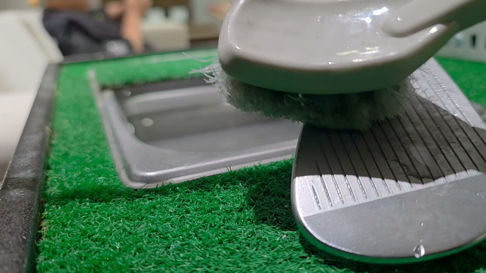 A golf wedge being brushed clean over a washing basin set into an artificial turf mat, the thick sole and the sharp leading edge both visible against the light.