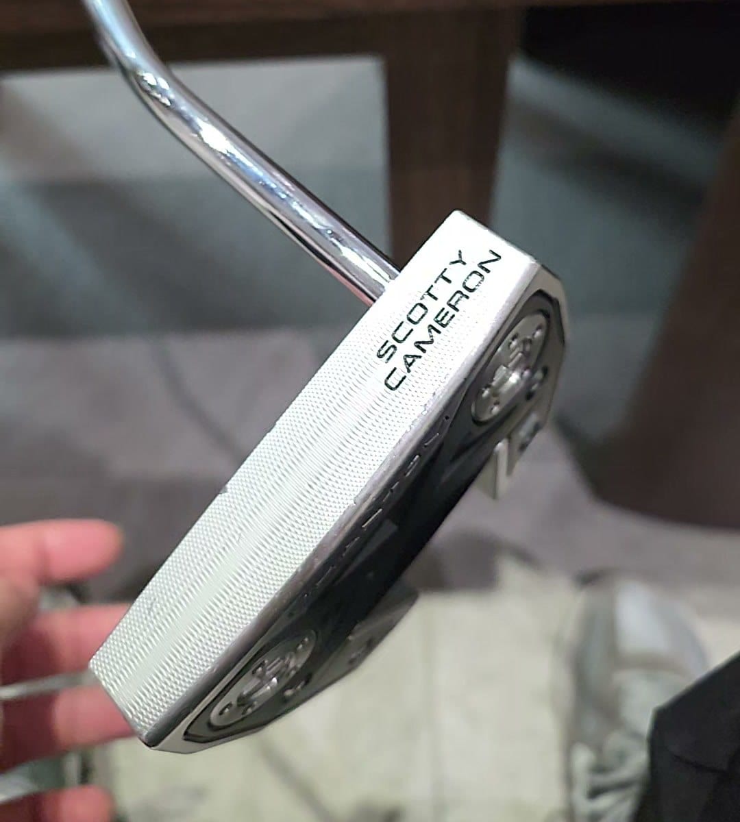A Scotty Cameron Phantom X 5 held up in one hand, the milled face and SCOTTY CAMERON stamp toward the camera with the two round sole weights tilted into view, and the shaft bending once above the head before it meets the heel end.